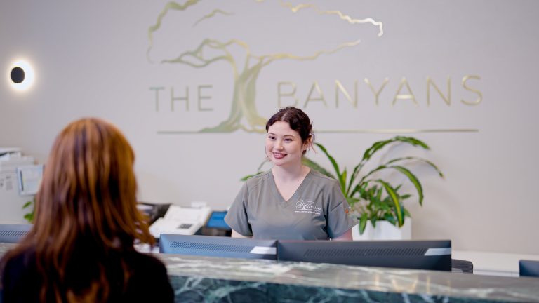 A smiling receptionist in grey scrubs stands behind a marble desk, welcoming a woman whose back is to the camera. The logo "THE BANYANS" is visible on the wall behind them.