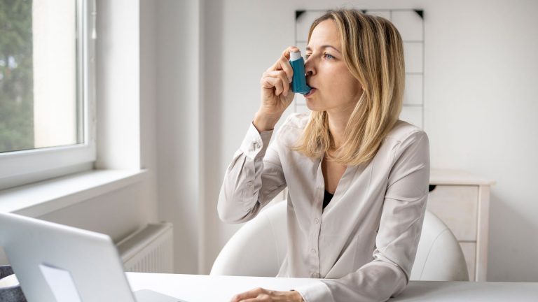 A woman sits at a desk using a laptop while taking medication with a blue asthma inhaler. She is seated in a bright, modern office with natural light coming through a nearby window.